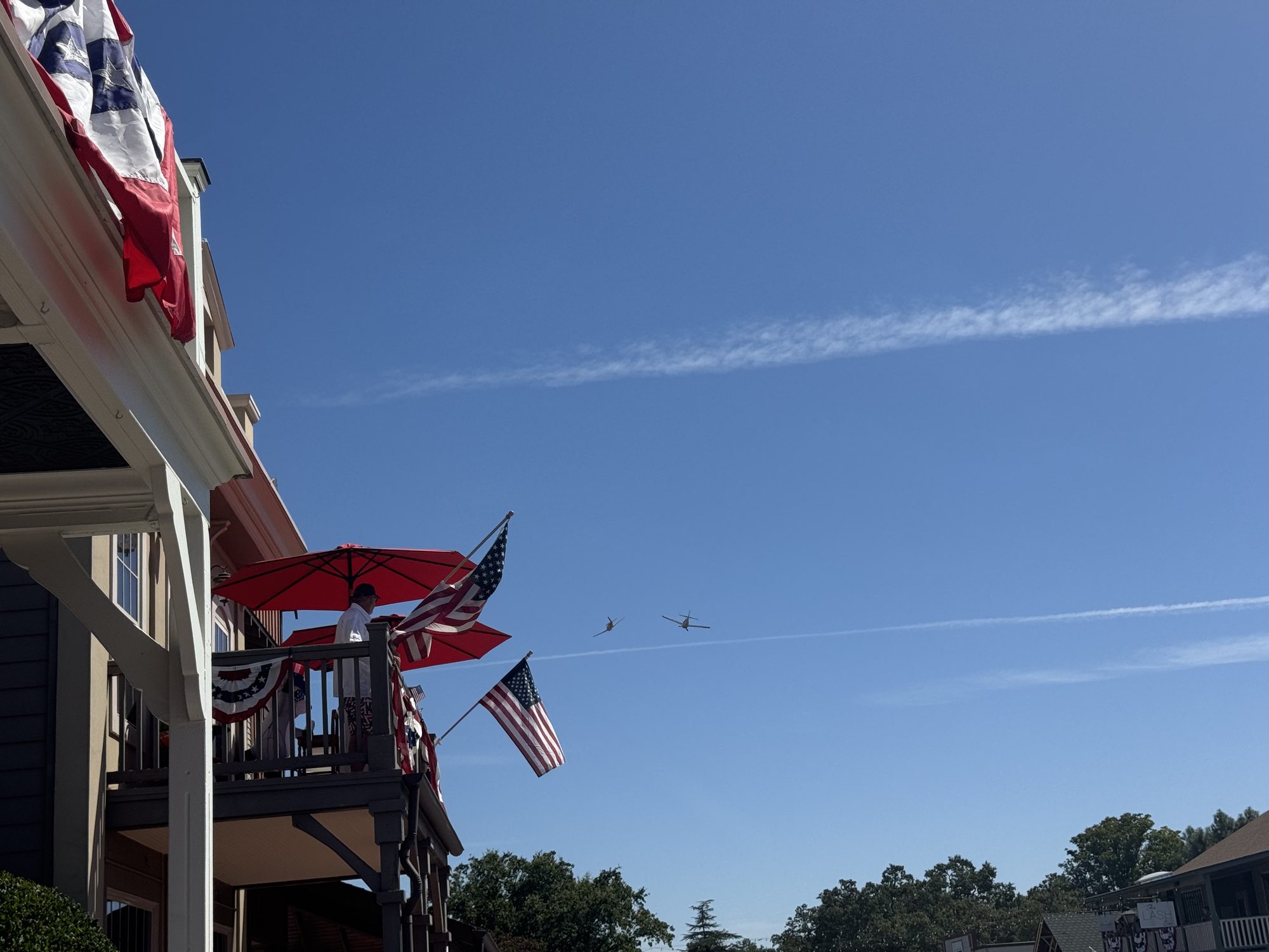 Two vintage Navy trainers making a low pass over a decorated balcony — American flags, red patio umbrellas, and bunting in the foreground against a clear blue sky