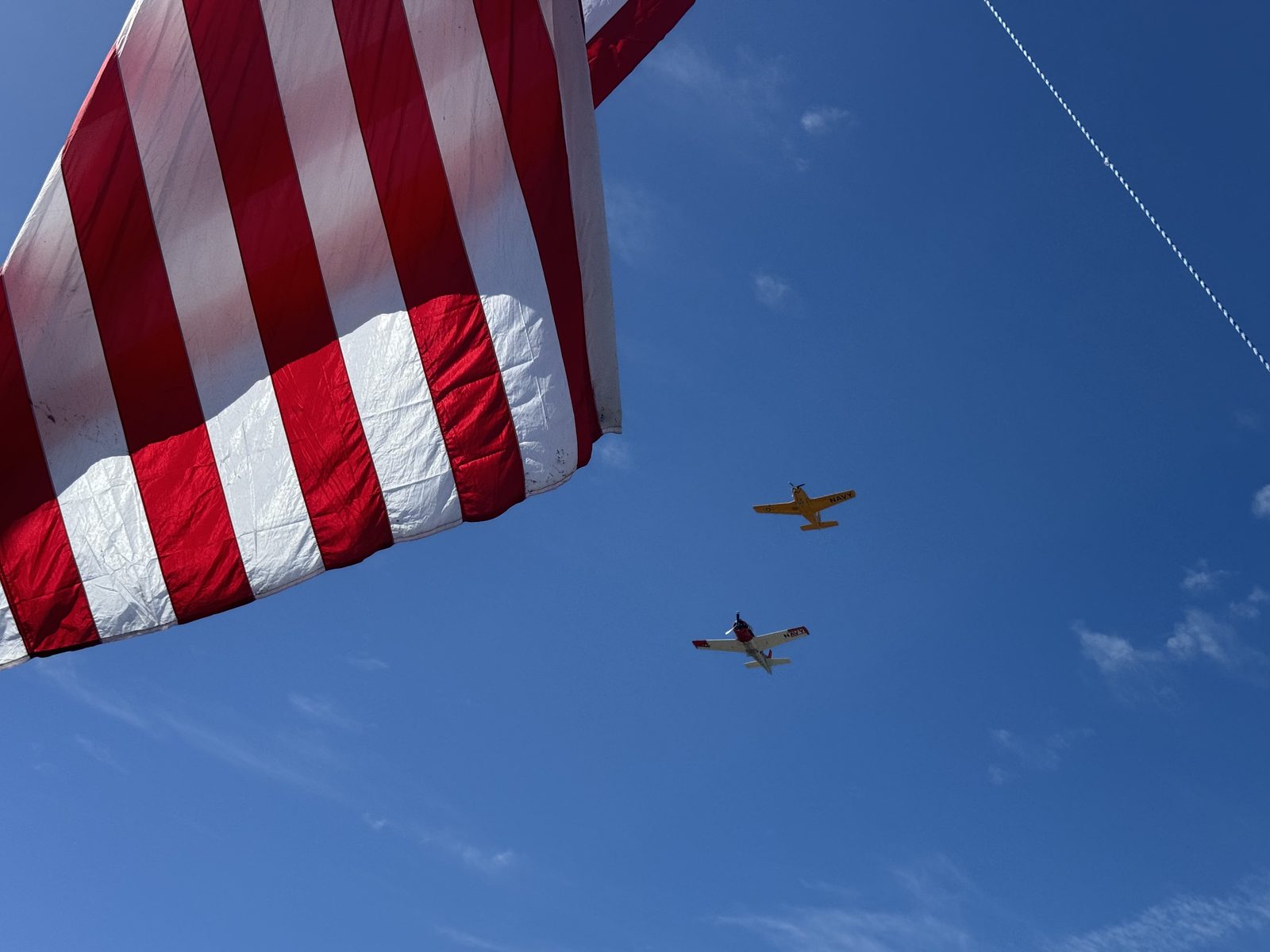 Two propeller warbirds banking through a clear sky, framed beneath the hanging stripes of the giant flag