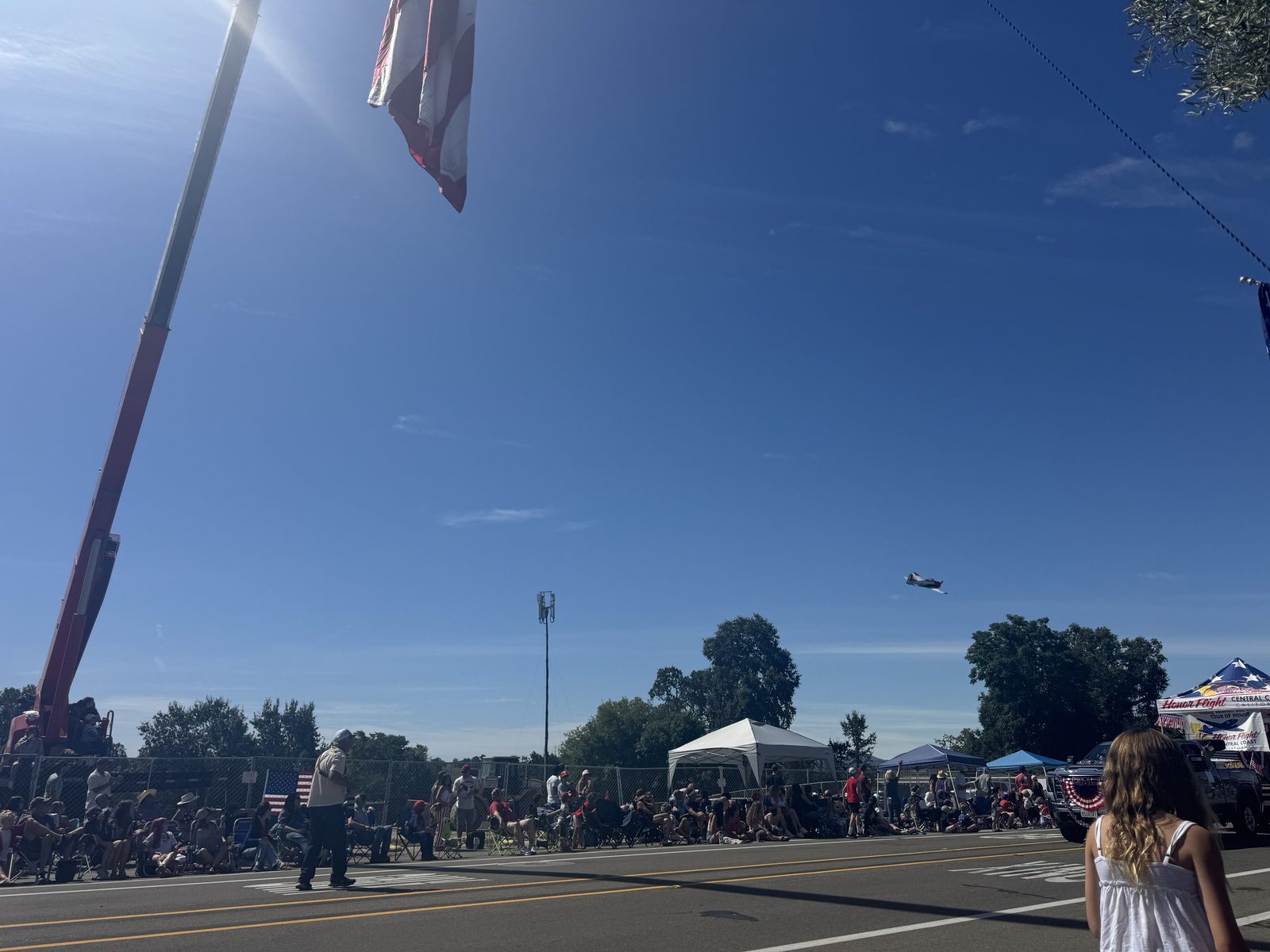 The parade crowd lining the street in lawn chairs under a bright sky, an Honor Flight tent along the route and a warbird overhead
