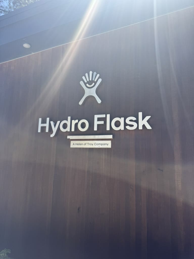 Hydro Flask headquarters sign — clean white logo on a dark wood-panel wall, "A Helen of Troy Company" plaque underneath