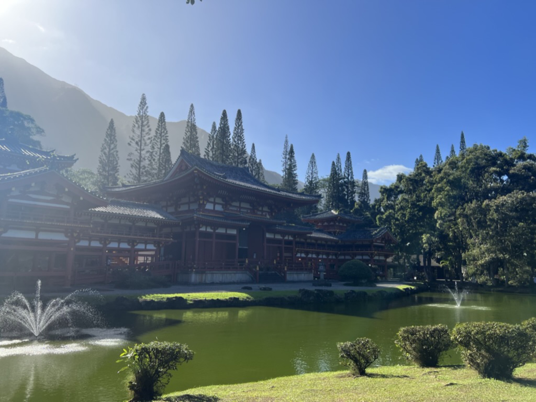 Byodo-In Temple in golden afternoon light — red pavilions, fountains in the green pond, mountains rising behind