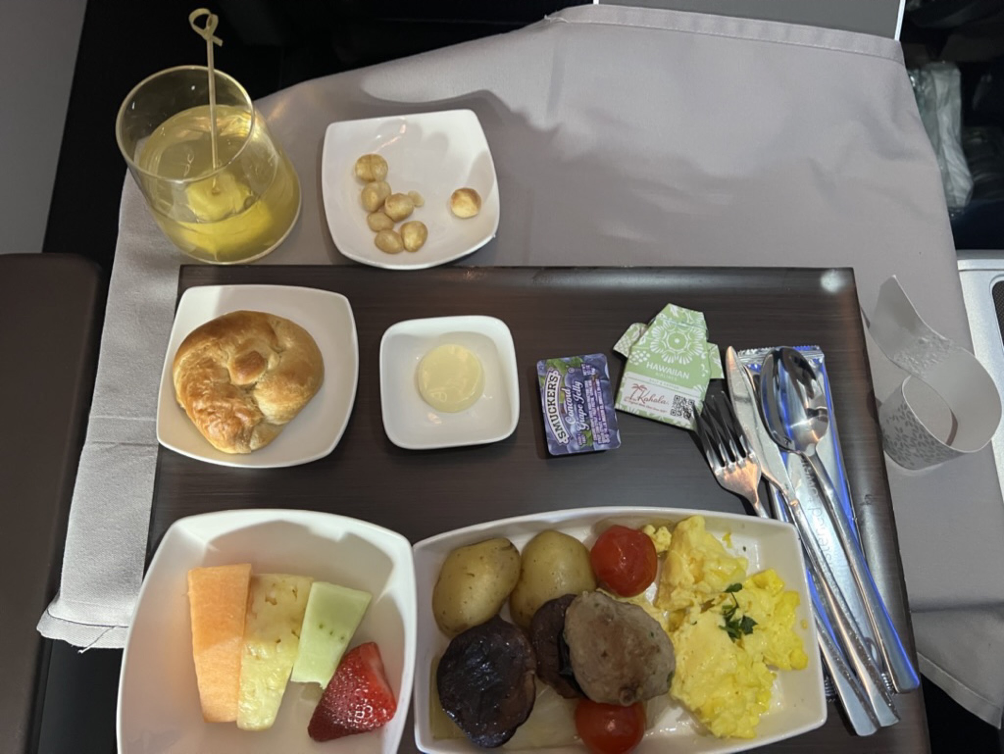 First-class breakfast tray on Hawaiian Airlines — fruit, scrambled eggs, sausage, a warm bun, macadamia nuts, butter, jam, and a tall Mai Tai in a clear glass