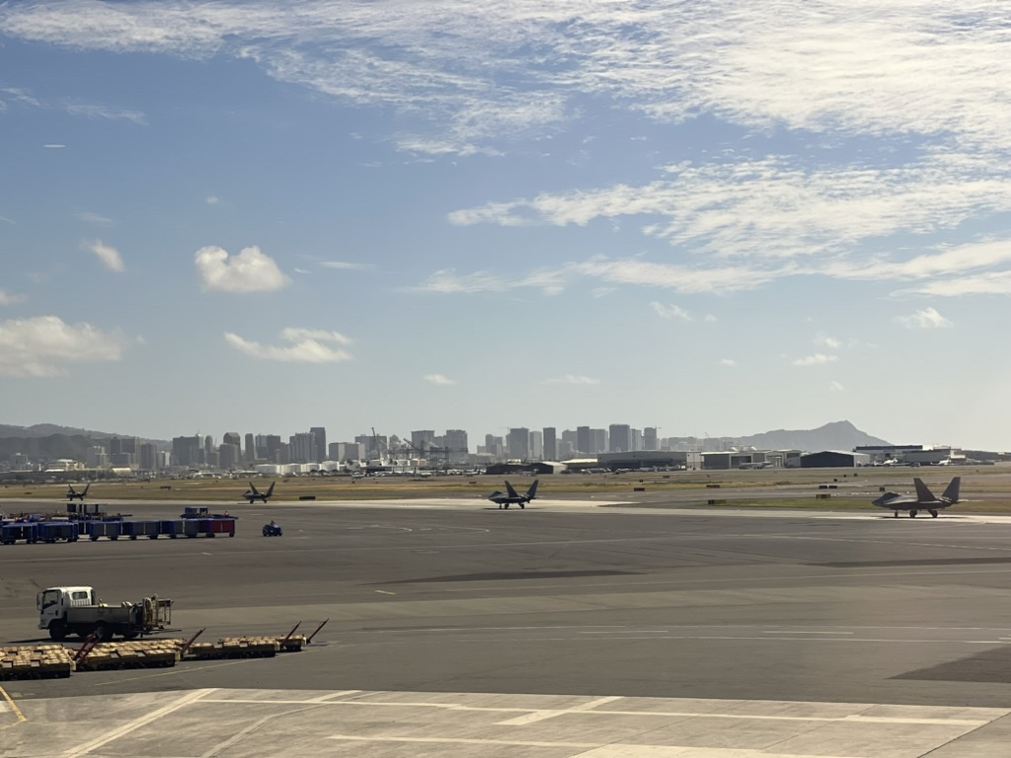 View from the airport observation area — Honolulu skyline in the haze, F/A-18 fighters lined up on the joint-base tarmac, Diamond Head's profile rising at the far end of the coast