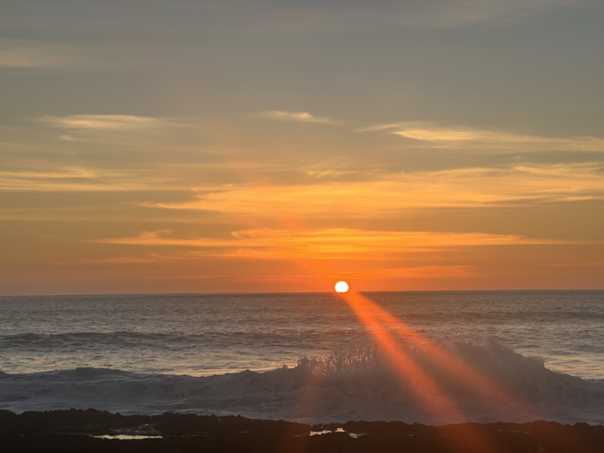 The sun touching the horizon as a wave breaks across the foreground rocks, the spray catching the orange light