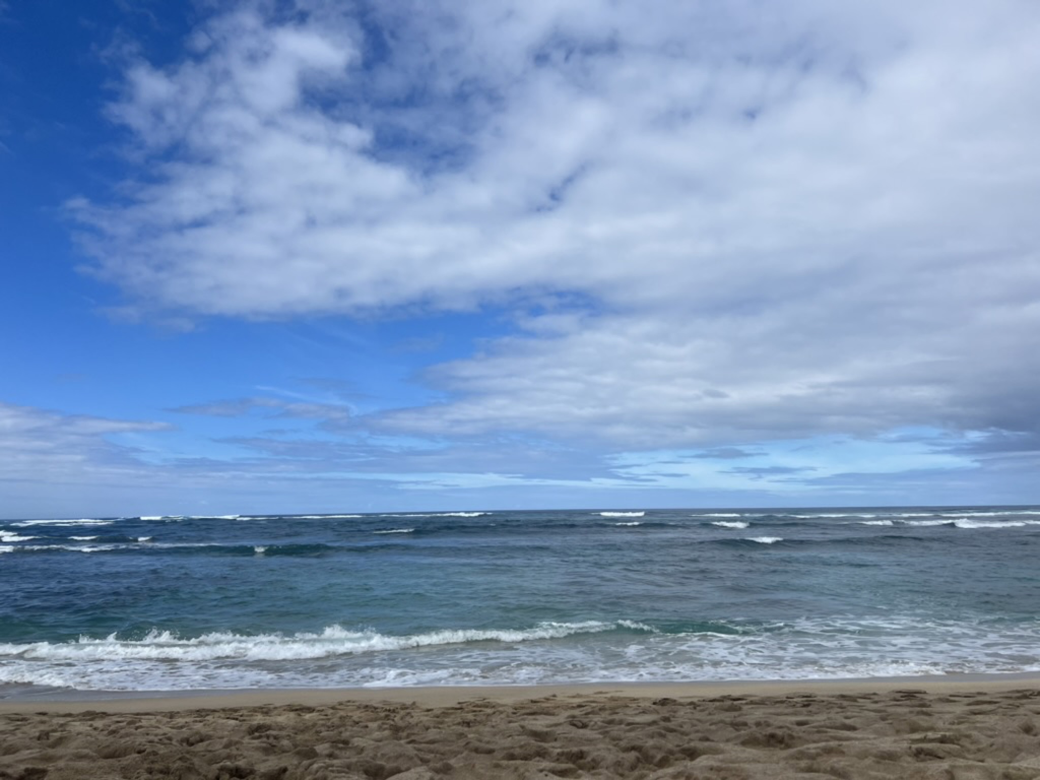 A North Shore beach — wide pale sand, breaking waves, scattered cumulus over the Pacific