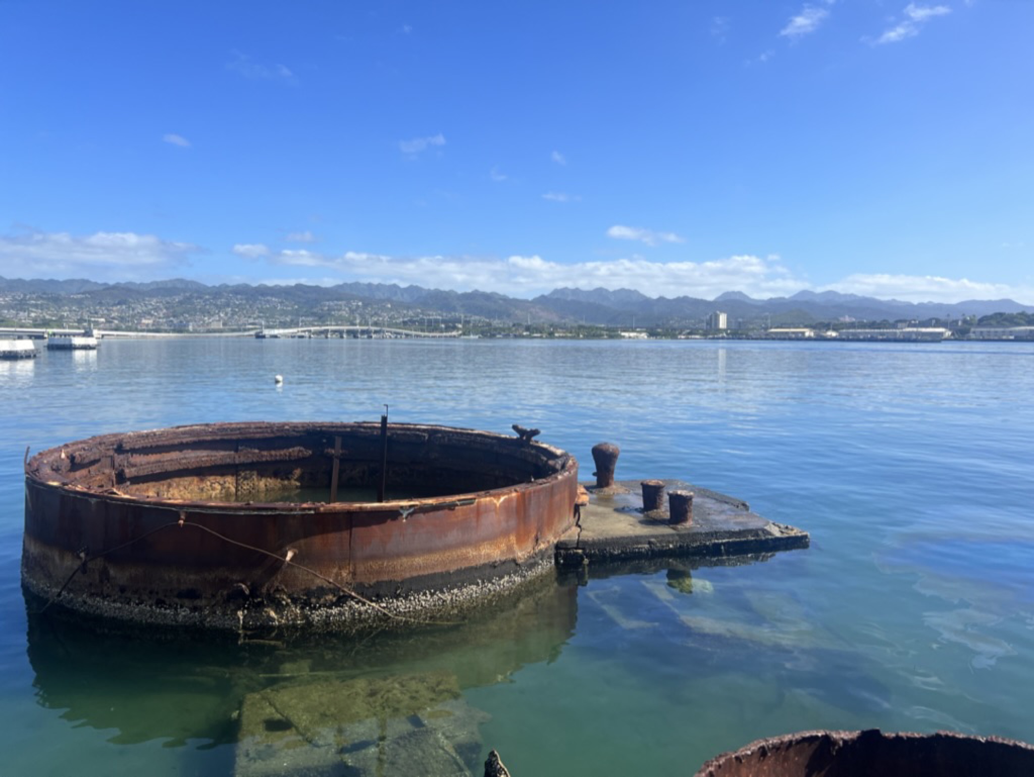 A rusted gun turret of the USS Arizona breaking the surface of Pearl Harbor, the Honolulu skyline visible across the water