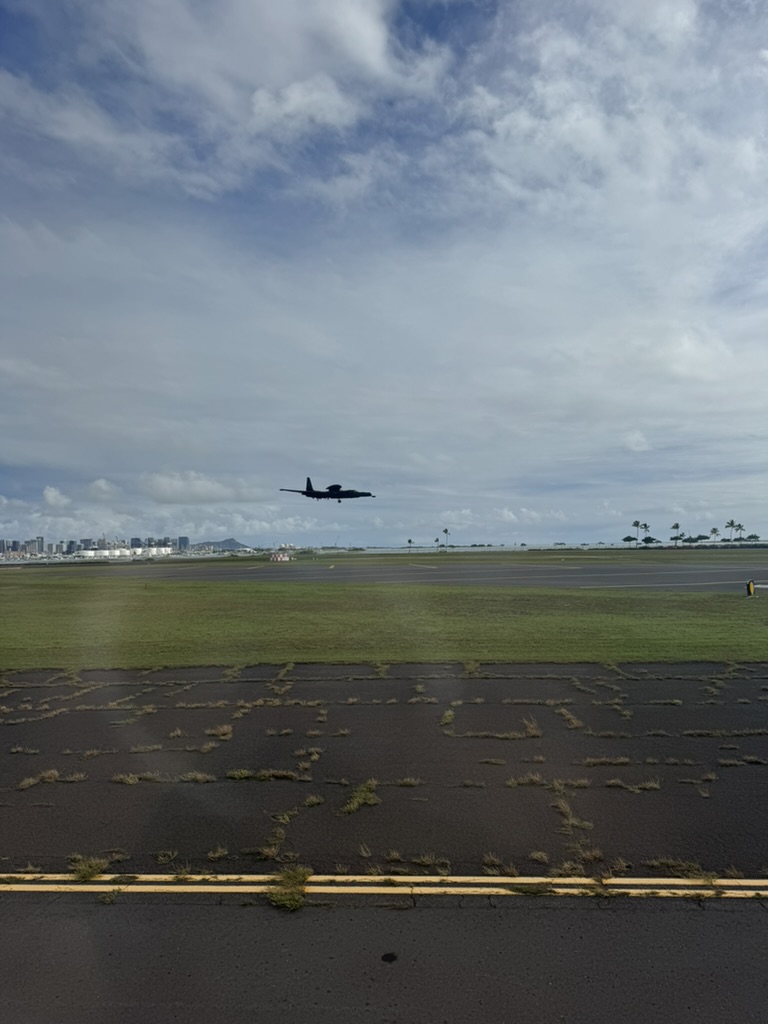 A US Navy E-2 Hawkeye aircraft on approach over the Honolulu airport runway — the Honolulu skyline and Diamond Head's profile visible across the field, scattered cumulus over the Pacific behind