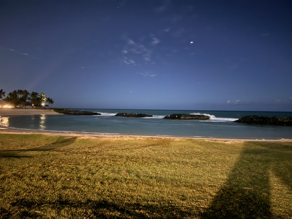 The Ko Olina lagoon at full dark with a bright moon high overhead — a single planet visible to the right, palm trees uplit at the far end of the bay, low surf rolling in
