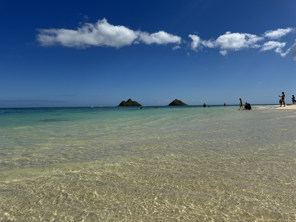 A wider Lanikai shot with the Mokes on the horizon, swimmers in the shallows, scattered cumulus over a deep-blue sky