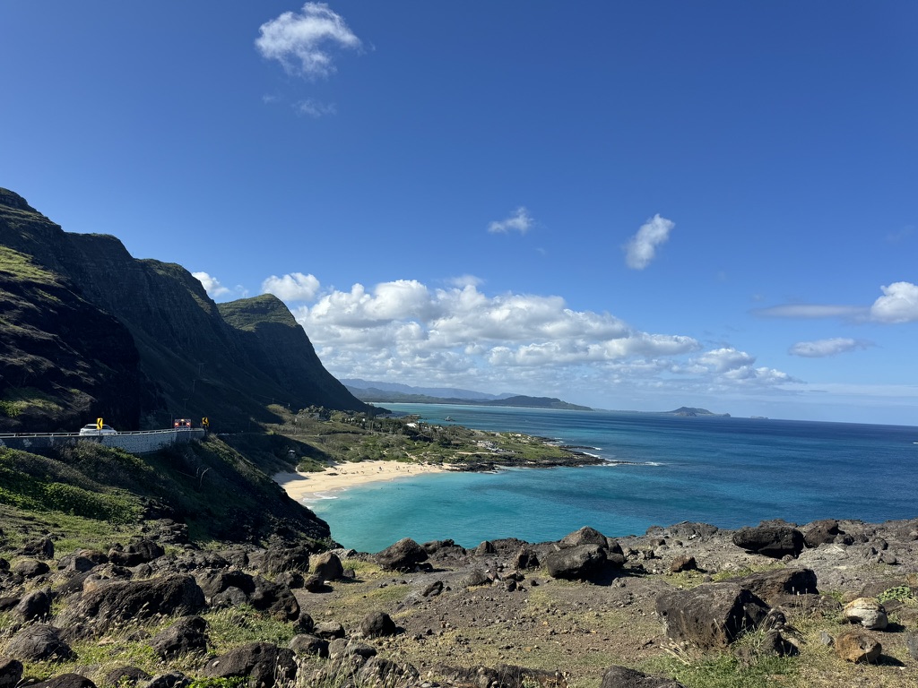 Panoramic view of Makapuʻu Bay from the lookout — sharp green cliffs at left, the curved sand beach below center, deep-blue Pacific stretching to the horizon