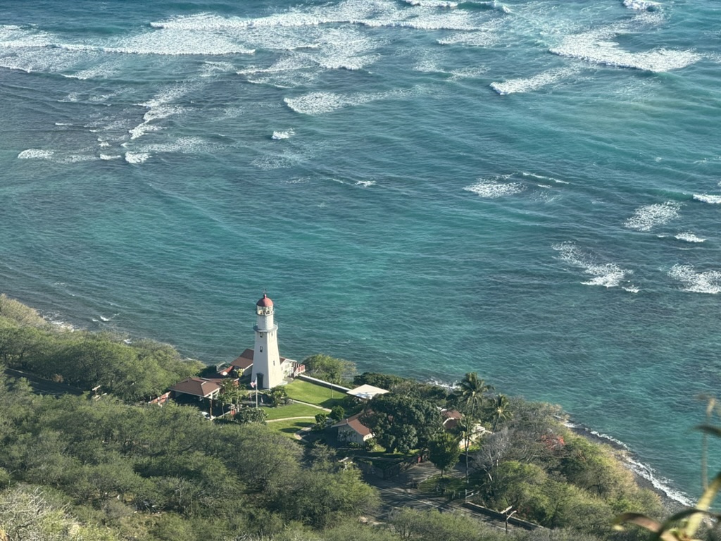 A wider take of Makapuʻu Beach from the cliff trail — the road snaking around the headland, the curved sandy bay below, the Pacific deep blue under scattered cumulus