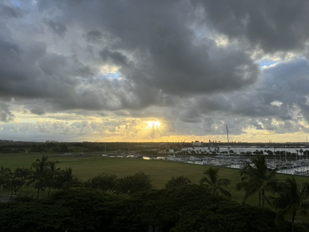 Dawn over the Kalaeloa marina from the room balcony — soft gold light breaking through a heavy cloud deck above the harbor cranes, sailboats and a green lawn in the foreground