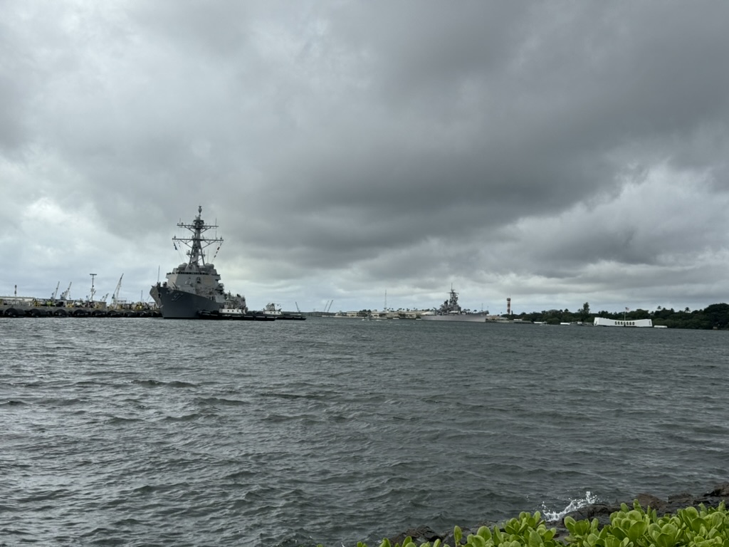 Wide view across Pearl Harbor — a docked destroyer at left, the USS Missouri mid-frame, the white USS Arizona Memorial at right under heavy overcast