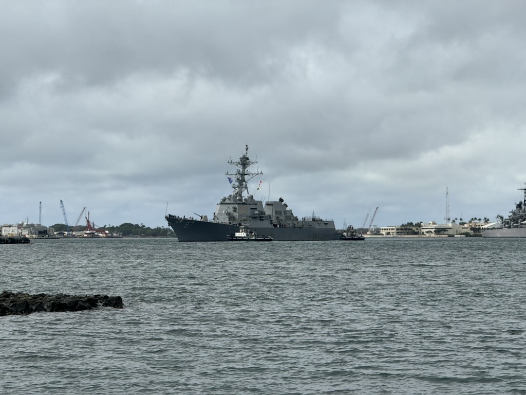 USS Halsey DDG-97 being moved across Pearl Harbor by two harbor tugs, an older docked vessel visible at the right edge of the frame