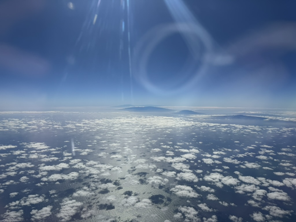 Two volcanic peaks of the Big Island silhouetted on the horizon through scattered cumulus, with a sun lens flare ringing the upper frame