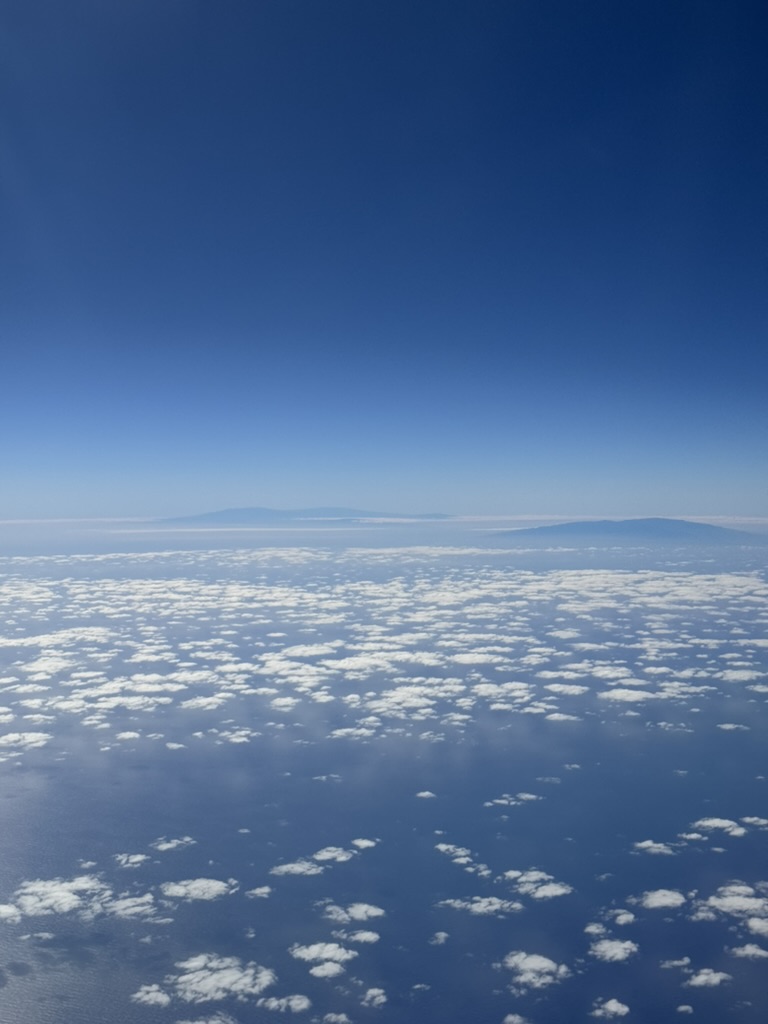 Vertical view from the airplane window — two distant island silhouettes on a hazy horizon, low cumulus dotting the Pacific below