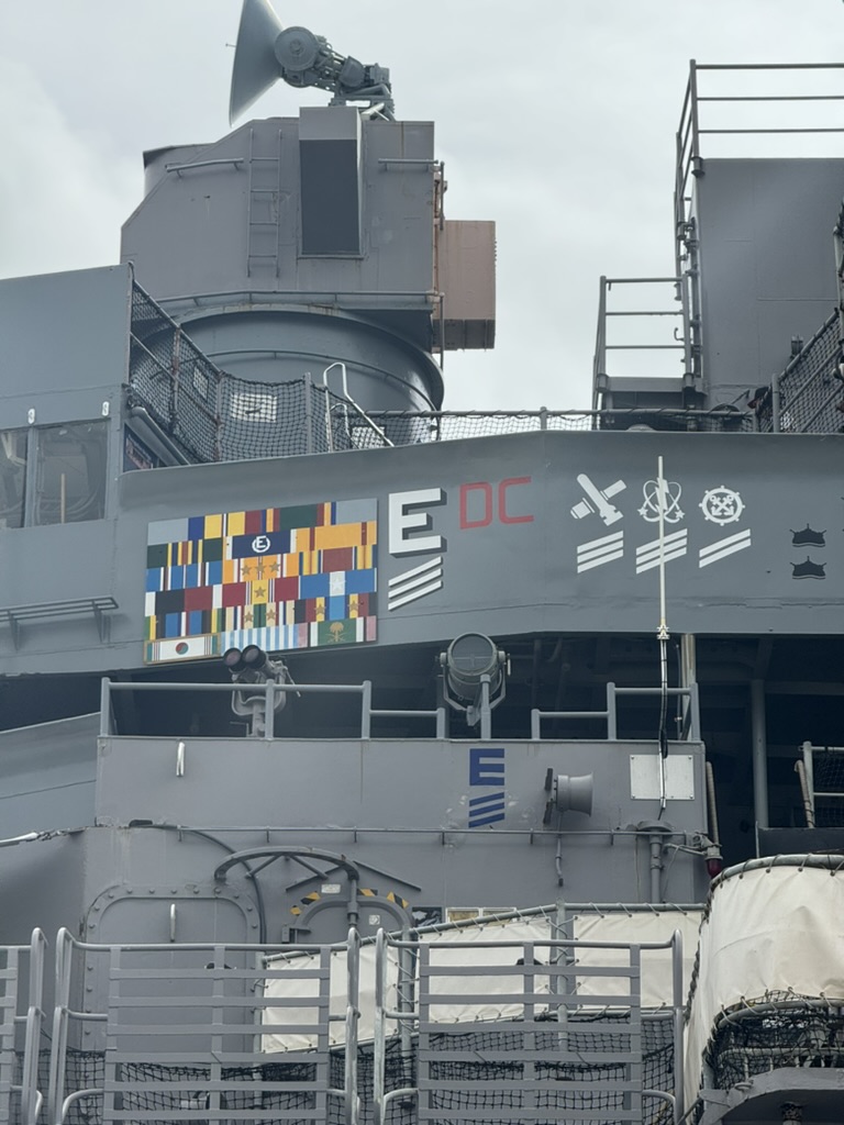 A square panel on the side of USS Missouri's superstructure displaying her battle awards — a grid of country flags representing combat tours, plus stylized icons for departmental awards
