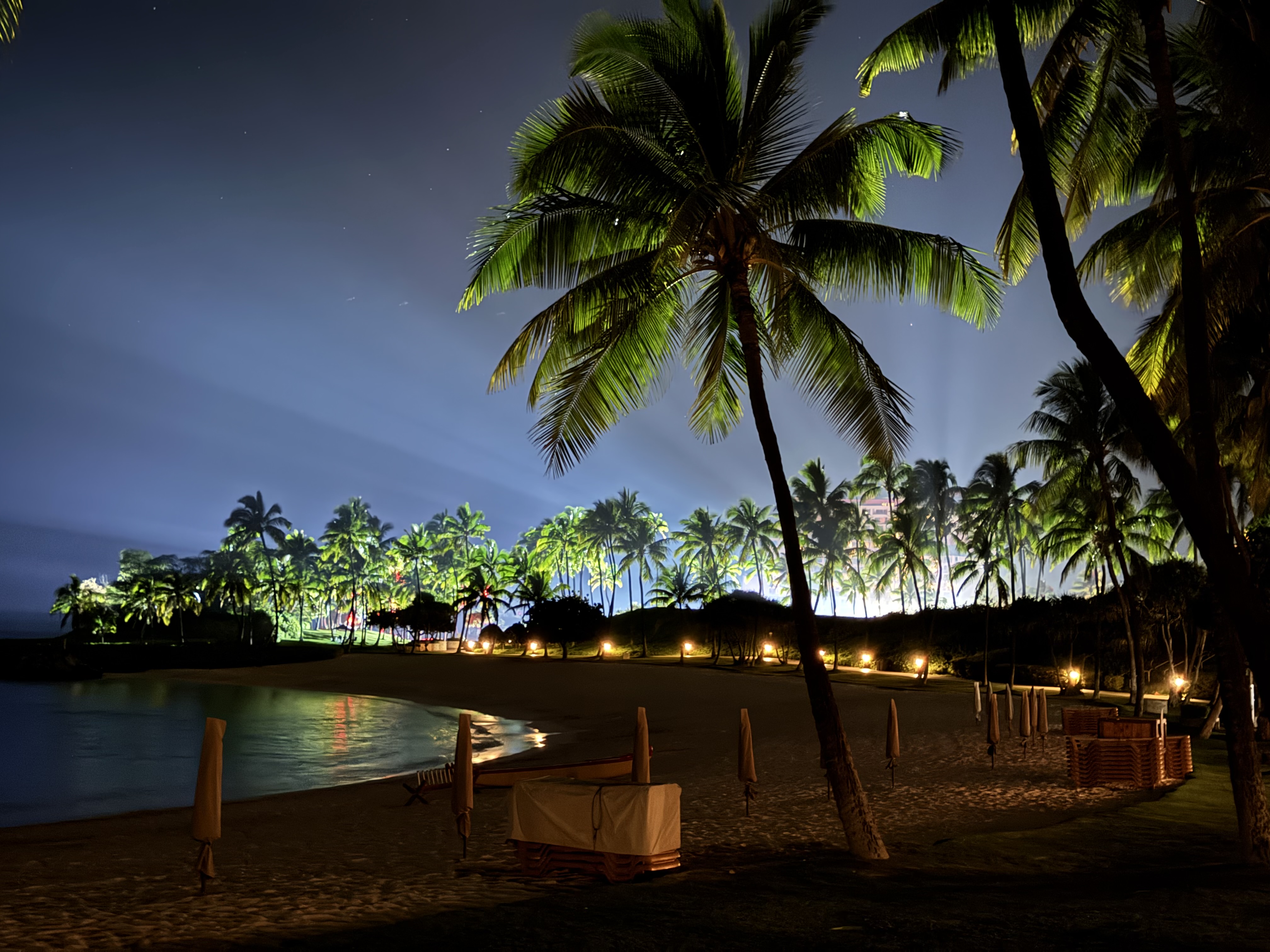 A wider take of the lagoon at night — palms uplit, beach cabanas tucked along the sand, distant resort lights bleeding through the trees