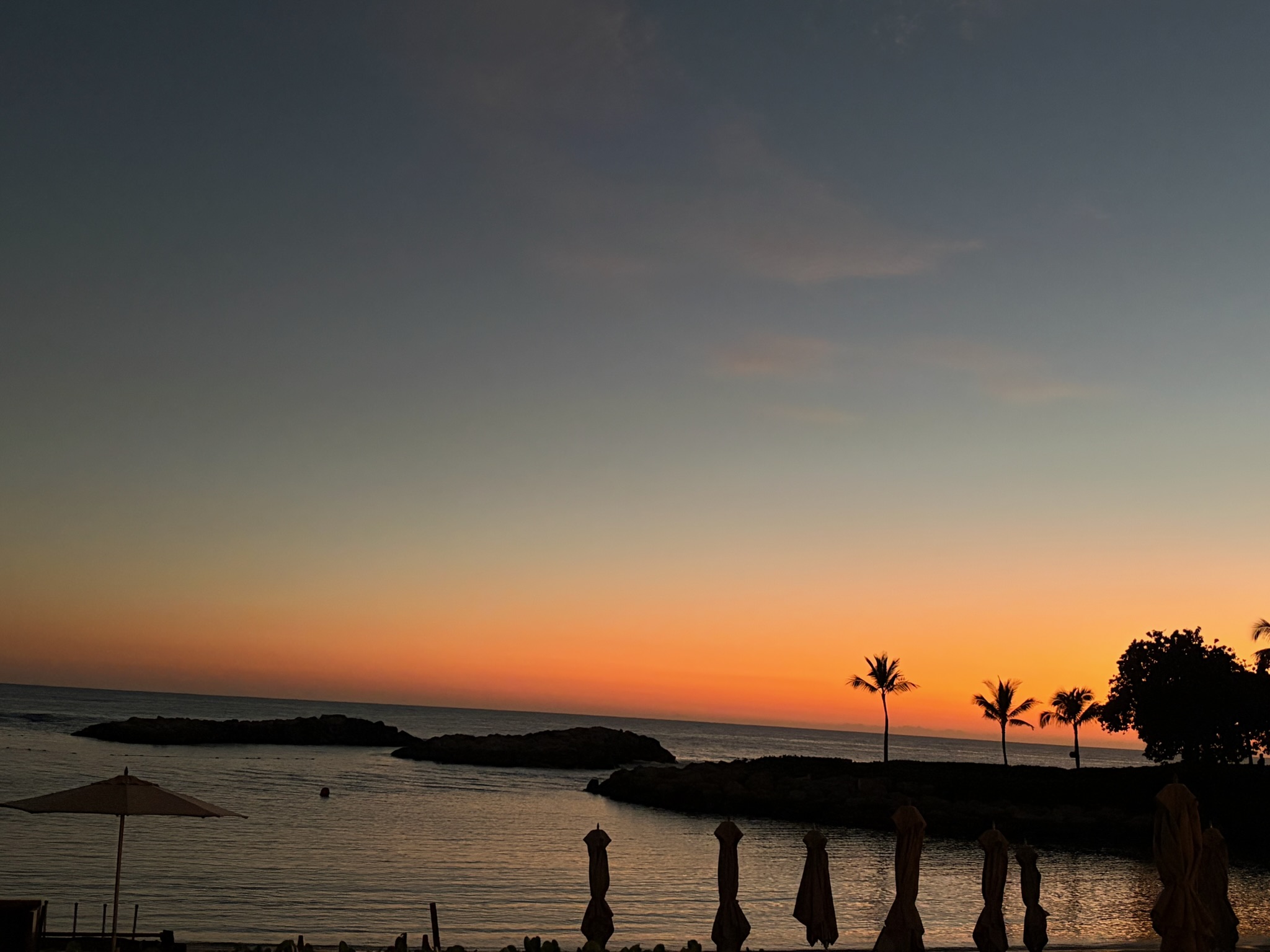 Sunset over the same lagoon, beach umbrellas tied up for the night, palms in silhouette