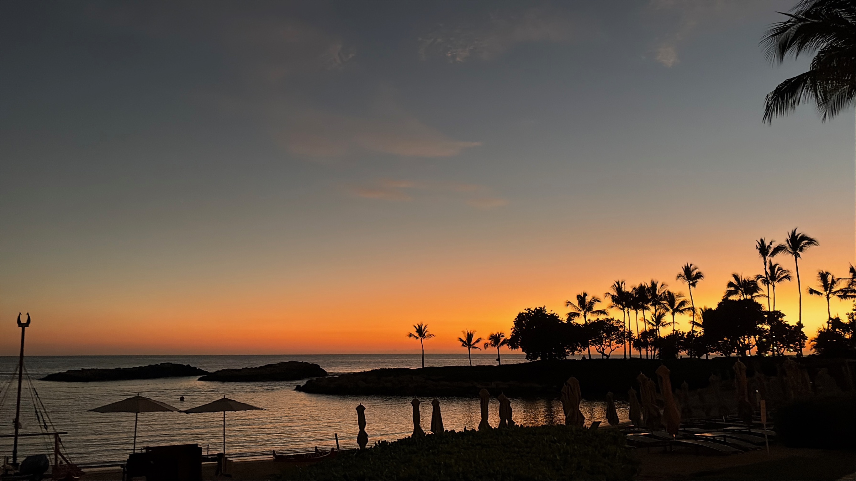 Wider sunset over the lagoon, beach umbrellas closed, palms standing against the orange band