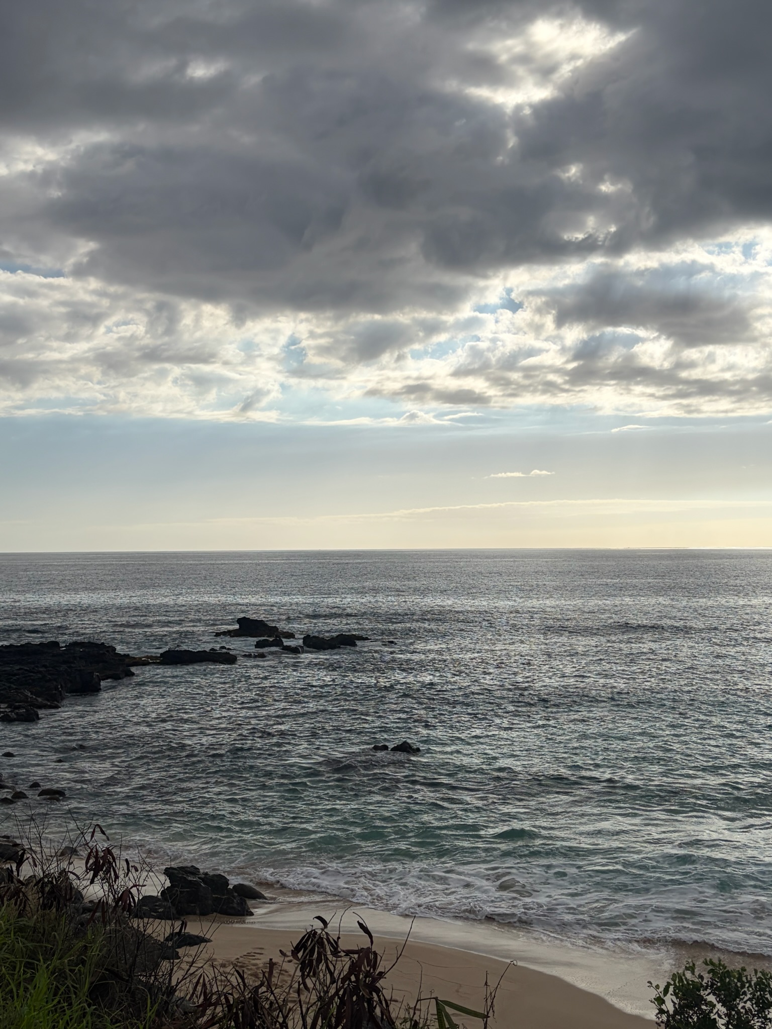 A wider stretch of windward coast under heavy overcast — moody clouds above, reef and rocks pushing into the surf below