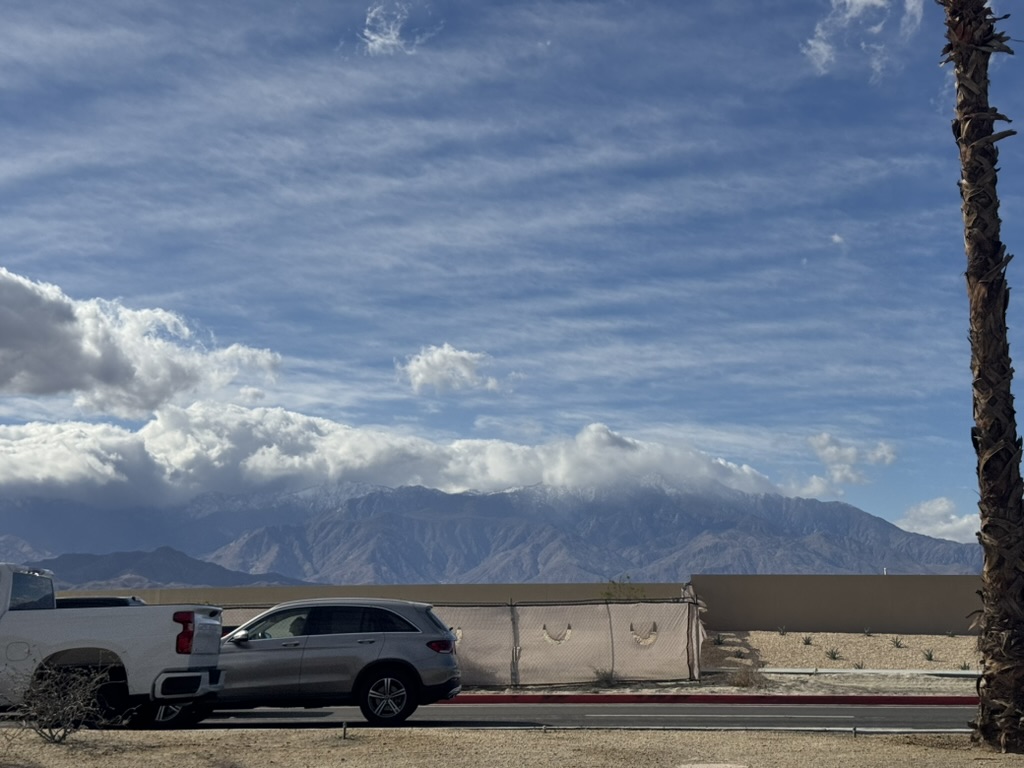Tesla parked roadside with the snow-dusted San Jacinto Mountains rising under a deck of clouds beyond